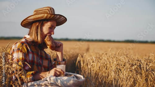 Young girl farmer in sun hat eats loaf of bread biting it drinking milk sits on wheat field with grain bag on knees. Harvesting, summer on farm, farmland, agribusiness, woman worker has break. photo