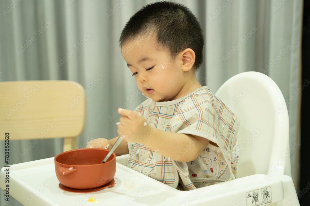 Baby boy eating with silver spoon by himself, dirty face and apron, sitting on white child chair at home Baby boy eating with silver spoon by himself, dirty face and apron, sitting on white child chair at home