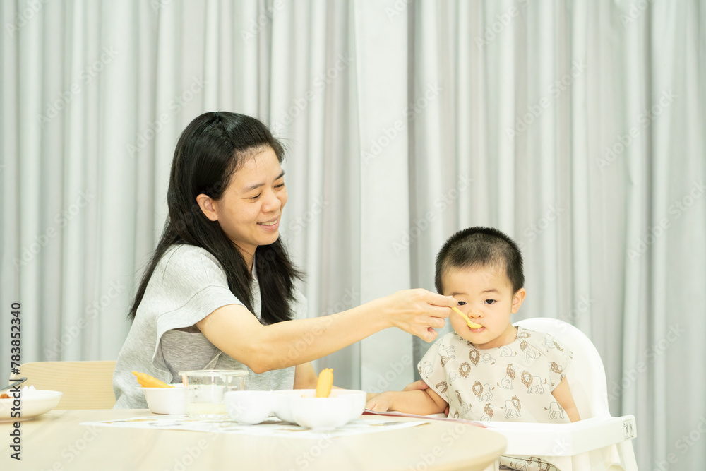 Asian mother feeding baby boy sitting on white highchair Asian mother feeding baby boy sitting on white highchair