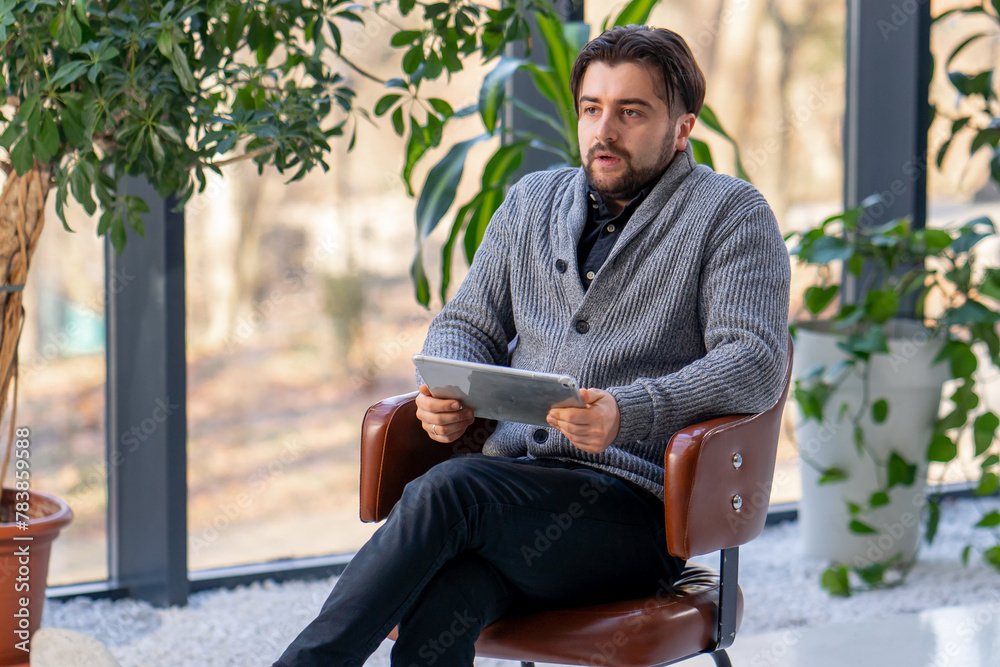 Fototapeta premium In a white hall young guy office worker on a brown chair works on a tablet