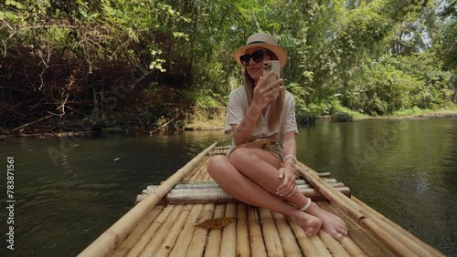 Female tourist floats on bamboo raft. Woman tourist enjoying the bamboo rafting on the river, films beautiful nature landscape on smartphone. Vacation, tropical tourism, forest adventure concept. photo