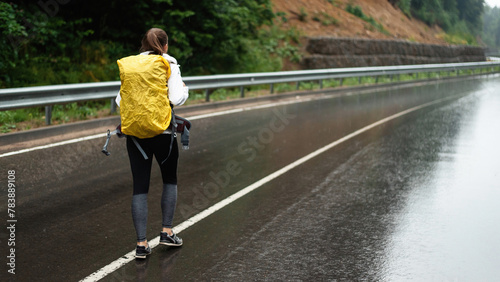 Back view of female hiker with yellow backpack walking on asphalt road with beautiful mountains around. Concept of people and leisure activity.