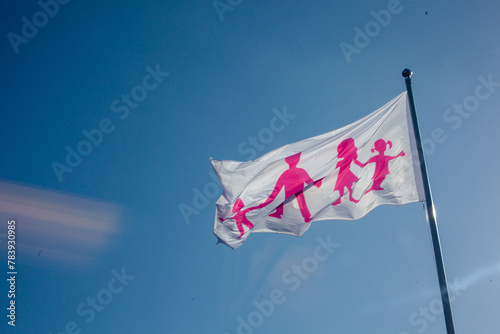 Flag where picture of parents with children. Pink sign on white flag. Blue sky background.