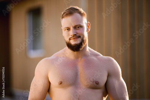 An athletic white man with beard stands alone near wooden rural house outdoors on summer evening in countryside.