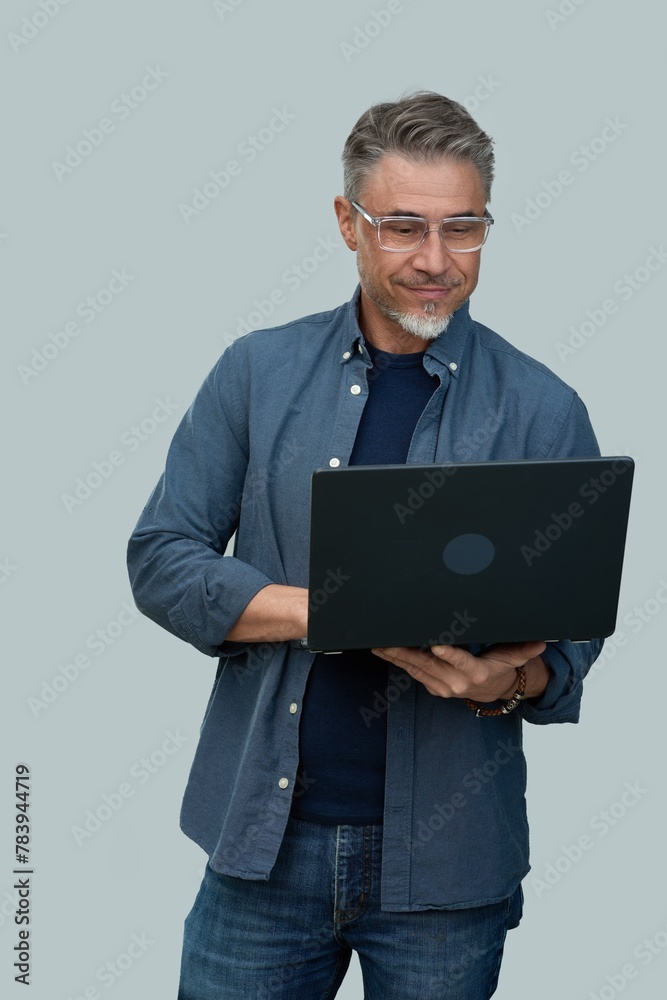 Fototapeta premium Casual mid adult man standing holding laptop computer. Portrait of happy middle aged male in 50s with gray hair and glasses, smiling. Isolated on white background.