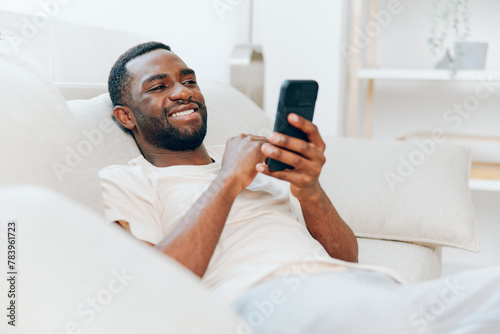 African American man resting on a black sofa, happily using his mobile phone indoors With a relaxed and confident demeanor, he types out messages, enjoying the convenience and connection of modern