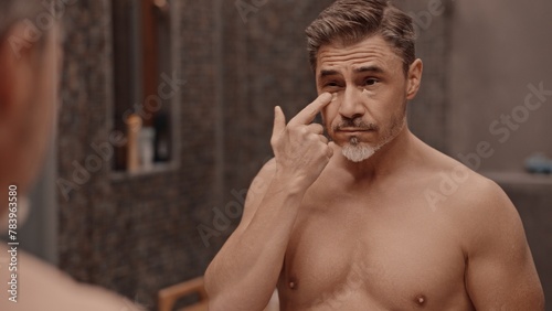 Shirtless mature adult man applying cosmetics in bathroom mirror. Using anti-aging eye cream for wrinkles to stop aging.