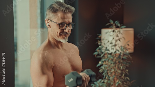 Fit, older shirtless man at home doing workout training exercise in morning by window, showcasing health and vitality, using barbell. photo