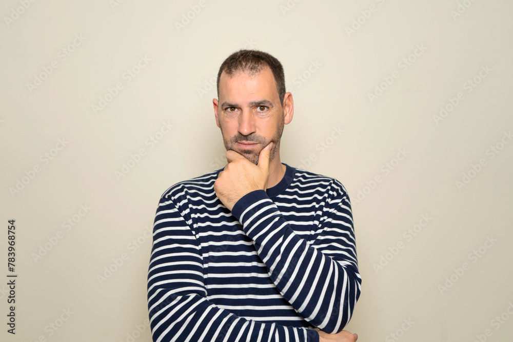 Hispanic man with beard about 40 years old wearing striped sweater thinking concentrated in doubt with finger on chin and looking at the camera wondering, isolated over beige background. Hispanic man with beard about 40 years old wearing striped sweater thinking concentrated in doubt with finger on chin and looking at the camera wondering, isolated over beige background.
