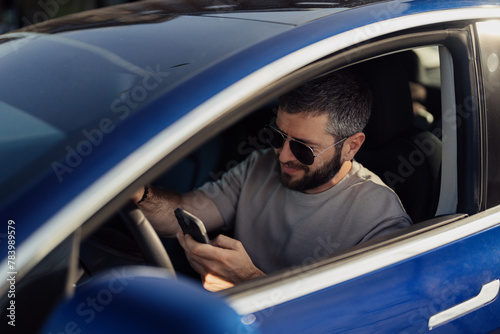 Smiling, bearded man wearing sunglasses interacts with his smartphone while sitting in the driver's seat of a modern electric car, showcasing a blend of technology and eco-friendly transportation. © arthurhidden