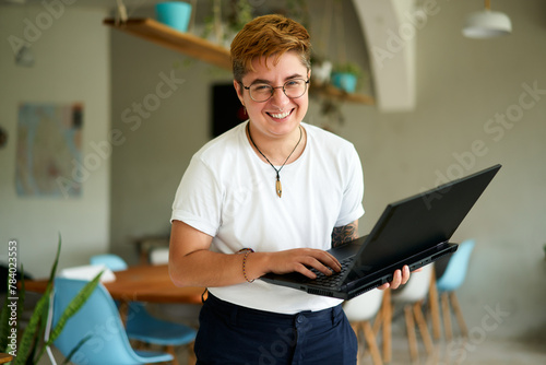 Smiling transgender professional holds laptop, works happily in modern office space. Inclusive workplace, young trans person enjoys career, experiences job fulfillment with tech in eco-friendly area. photo