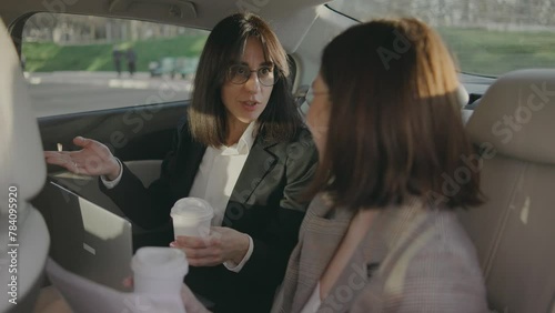 Two beautiful business women are sitting in the back seat of a car and solving business projects drink coffee and laugh in their free time. Business lady in the back seat of a luxury taxi with a white