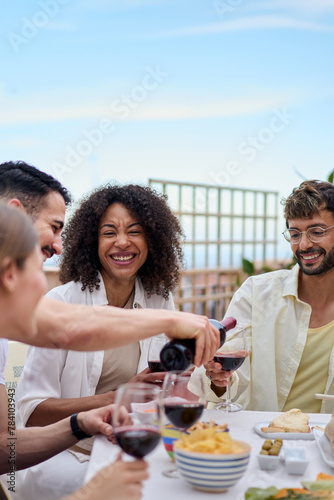 Vertical. Young smiling man serving red wine to guests at food table in celebration with happy friends on rooftop. Group of cheerful multiracial friends gathered for lunch party on outdoor terrace