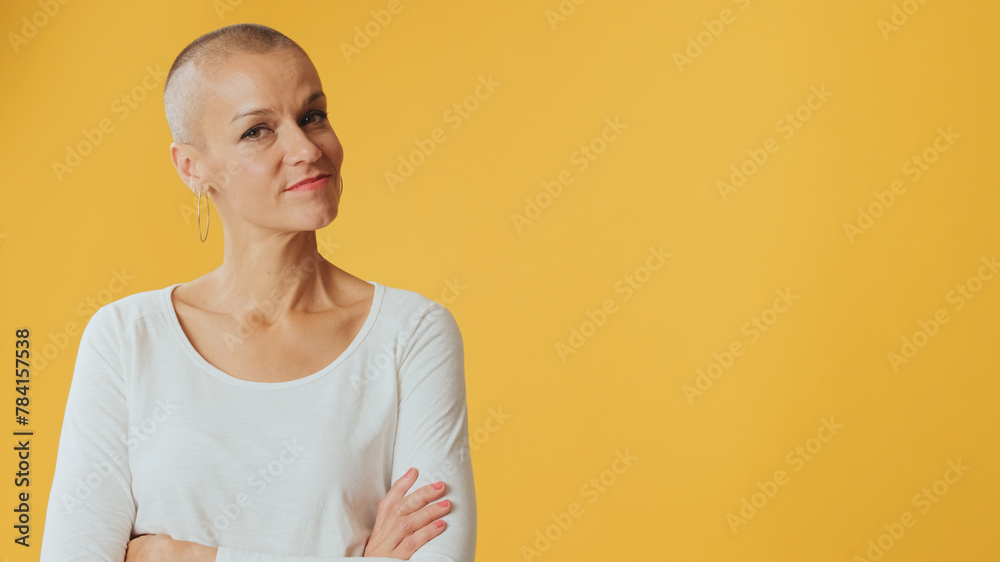Young hairless woman crossing her arms and looking at camera with smile, advertising space, isolated on yellow background in studio Young hairless woman crossing her arms and looking at camera with smile, advertising space, isolated on yellow background in studio