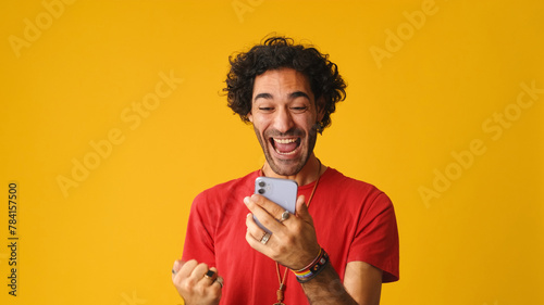 Surprised man with curly hair, dressed in red T-shirt,  reading good news on mobile phone celebrates victory isolated on yellow background in studio