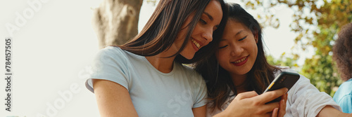 Young multi ethnic smiling women are watching videos on their phone. Girls laugh at picnic on summer day outdoors, Panorama