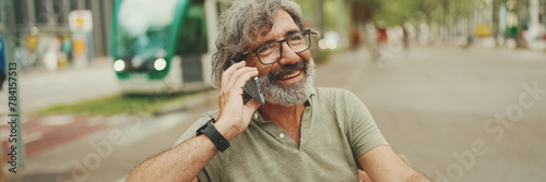 Laughing middle-aged man with gray hair and beard wearing casual clothes sits on bench and uses mobile phone, Panorama. Mature gentleman in eyeglasses talking on smartphone