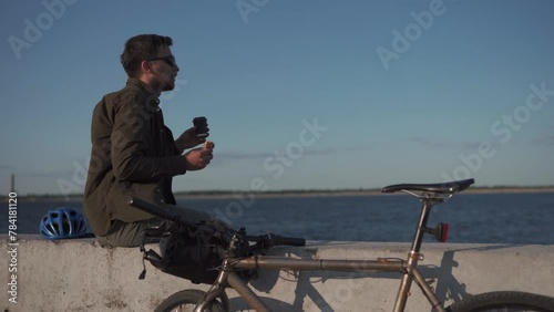 Cyclist sits on concrete fence near sea and drinks from disposable cup of coffee and eats sandwich on bicycle trip. Man has lunch after cycling near lake. Snack after work overlooking a large pond. photo