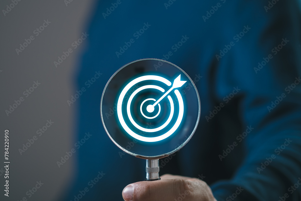 Hand holding magnifier glass with white dartboard with arrow for setup and focus in objective focus target goal concept.
Hand holding magnifier glass with white dartboard with arrow for setup and focus in objective focus target goal concept.