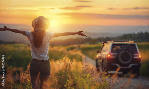 A woman is standing on the back of her car with arms outstretched, enjoying nature and freedom during vacation or travel trip at summer sunset time #784276983