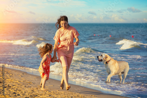 Young happy mother with little daughter holding hands and running barefoot on the beach. The dog runs nearby. Mother's Day concept