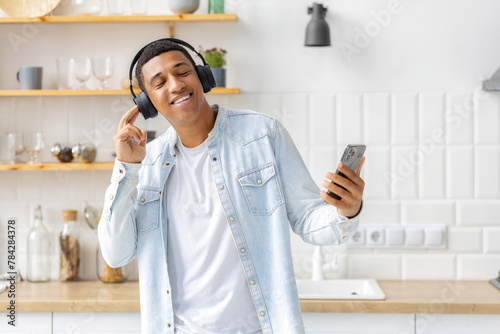 Happy positive African American man dancing and listening to music on headphones at home kitchen