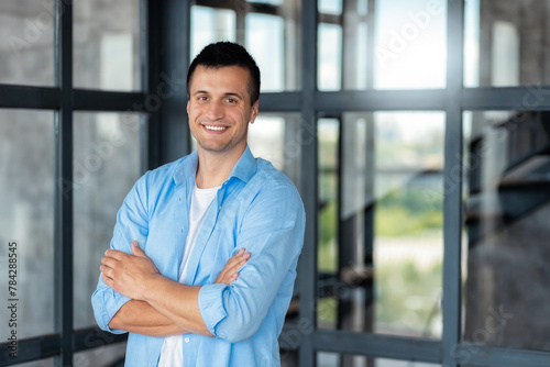 Young successful Caucasian man entrepreneur or an office worker stands with crossed arms in a modern office, looking at the camera and smiling