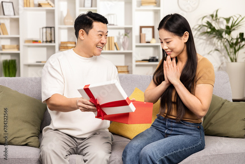 Surprised woman receiving a gift from a man, both sitting on sofa at home, expressing excitement and joy, with a cozy interior background © puhhha