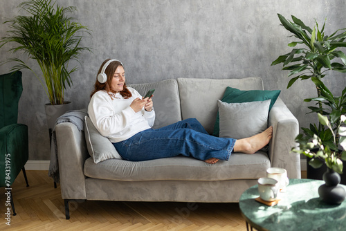Smiling girl relaxing at home, she is listening to music, using a smartphone and wearing white headphones. Young 30s woman hearing enjoying good audio sound lounge on couch at home, full body