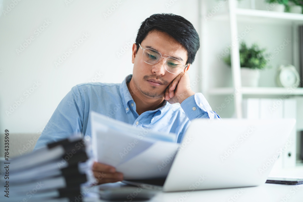 Fototapeta premium A man is sitting at a desk with a pile of papers in front of him. He is looking at a laptop and he is tired or stressed