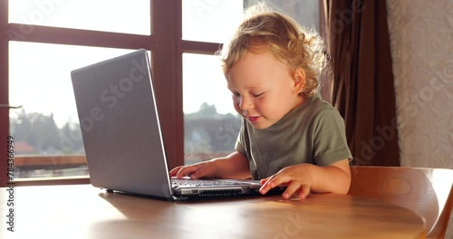 A young boy sits before a laptop, pressing keys and tilting his head to peer at the lettering. Little one imitating adults with cheerful curiosity, exploring the digital world with joy and wonder. photo