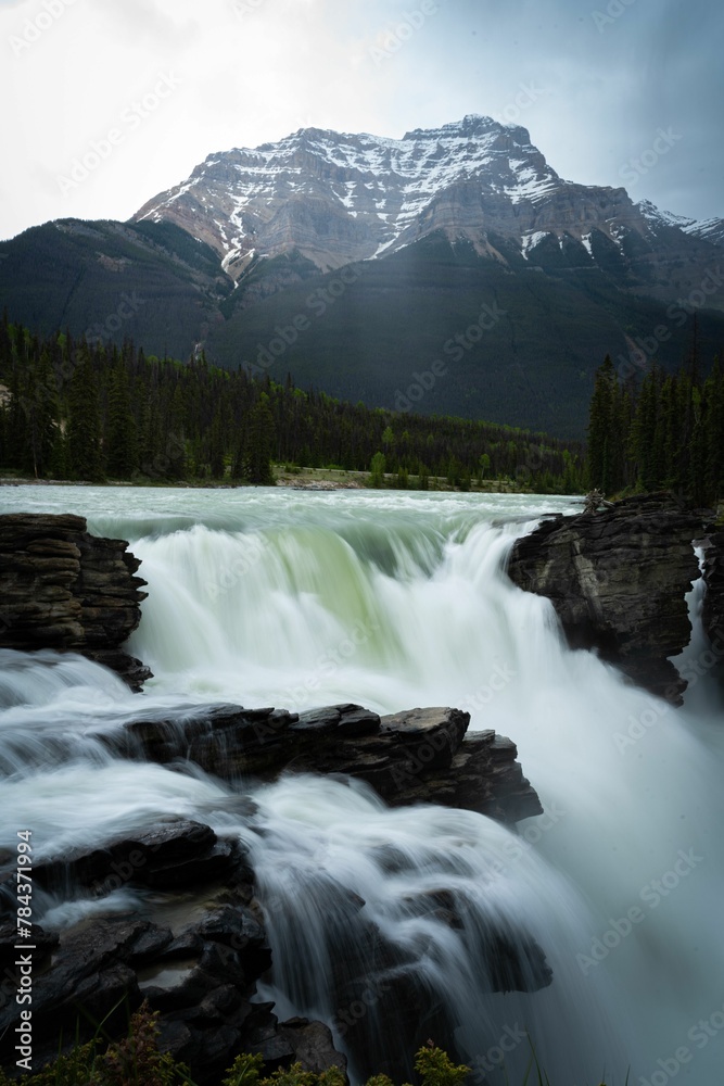 Long exposure of a foamy waterfall with forest and snowy mountains in the background Long exposure of a foamy waterfall with forest and snowy mountains in the background