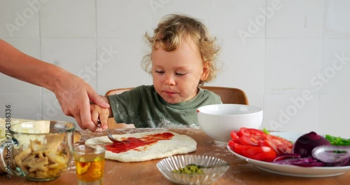 Young boy is learning to make homemade pizza, scooping tomato sauce with spoon, his mom takes his hand and shows him how to spread sauce on dough. Little one is more interested in tasting everything
