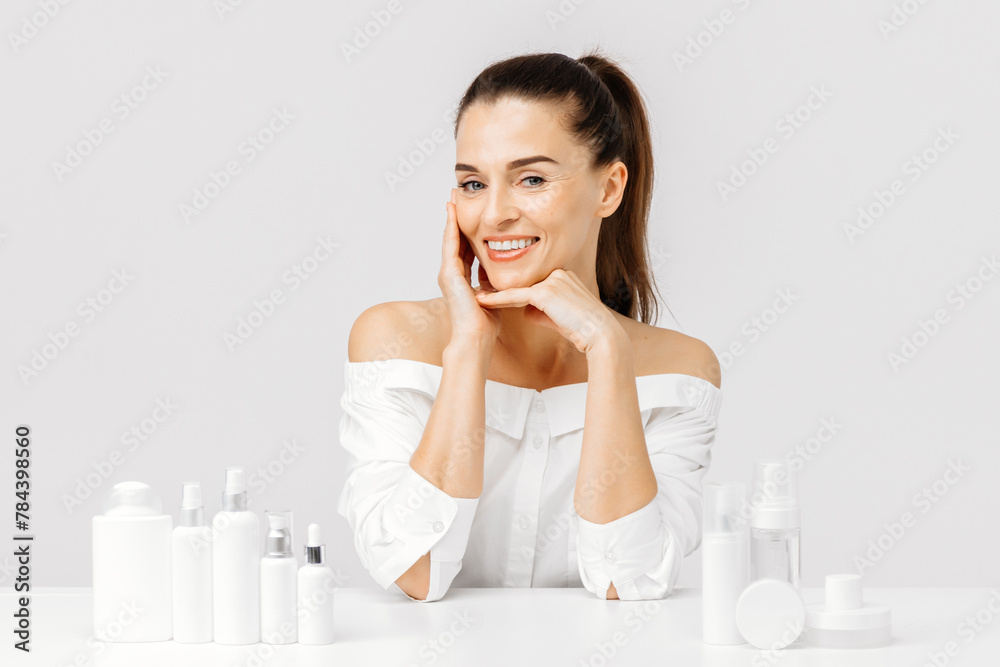 A happy mature middle-aged woman touches her face and looks at the camera among a mockup of cosmetic products. The concept of caring for healthy, dry skin, advertising of cosmetics and cosmetology. A happy mature middle-aged woman touches her face and looks at the camera among a mockup of cosmetic products. The concept of caring for healthy, dry skin, advertising of cosmetics and cosmetology.
