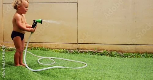 Young child turns on handheld water sprinkler, first spraying it onto ground before playfully directing stream at nearby puppy. Little French Bulldog joyfully shakes off water and run away. photo