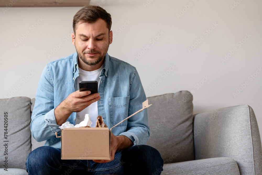 Man in a denim shirt unpacks a package and takes a photo of his purchase, internet order. Person sharing feedback with online store using his smartphone. Man in a denim shirt unpacks a package and takes a photo of his purchase, internet order. Person sharing feedback with online store using his smartphone.
