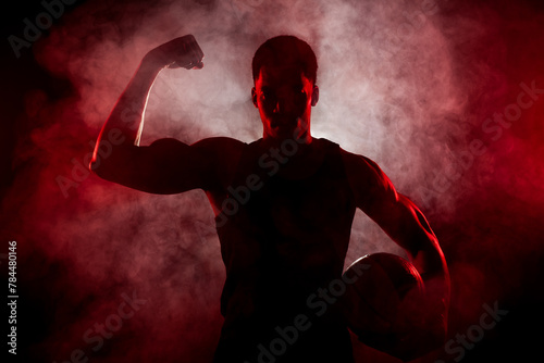 Silhouette basketball player side lit with red color holding a ball with smoke in the background. Serious african american man showing muscles
