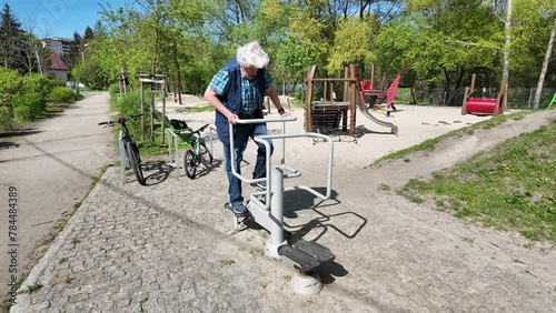 Old gray-headed senior 70-80 exercising in an outdoor creaking stepper machine in an open public garden gym park with a playground in Krakow, Poland. 4K video recorded with sound of the equipment photo