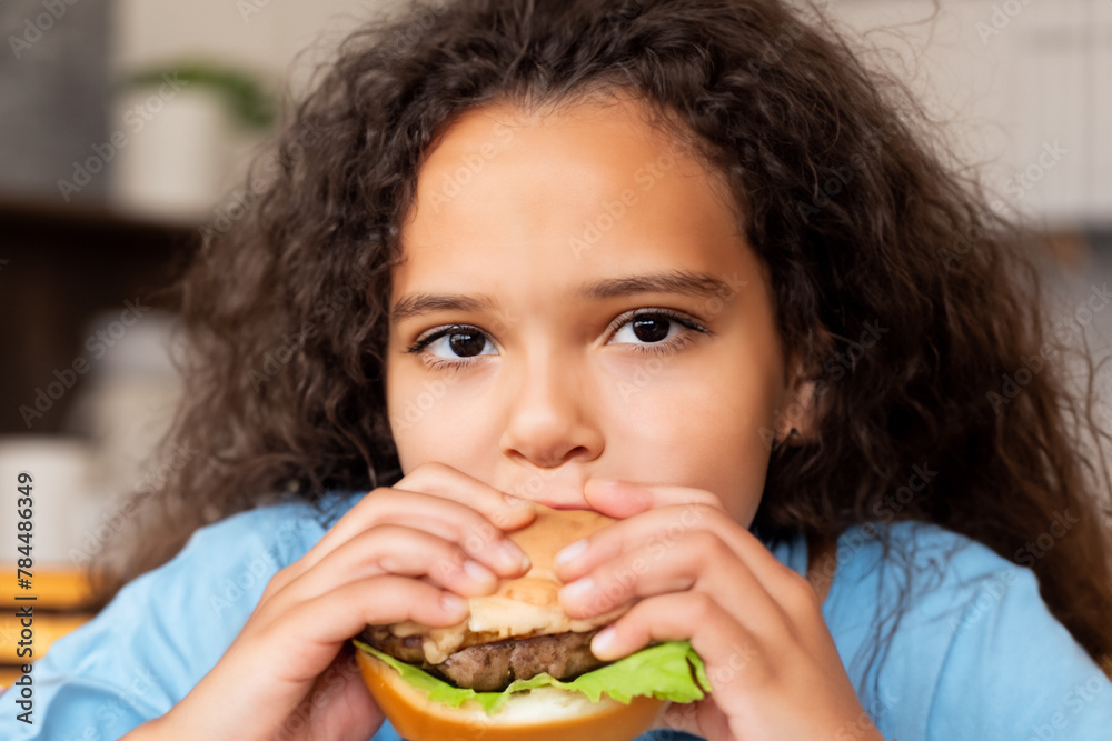 girl with a hamburger. little afro girl in a blue t-shirt sits at the table and eats a big hamburger, close-up, food concept girl with a hamburger. little afro girl in a blue t-shirt sits at the table and eats a big hamburger, close-up, food concept