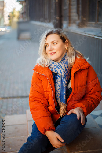 Happy smiling middle aged woman in orange down jacket sitting on concrete stairs outdoors