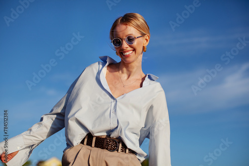 Contemporary happy woman in fancy clothes oversized shirt and sunglasses, joyfully smiling against blue sky. Trendy, happy, and stylish summer vibes photo