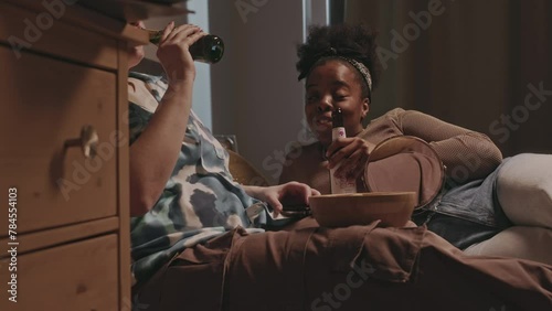Two multiethnic cheerful girlfriends lying on bed with cider bottles and gossiping during sleepover