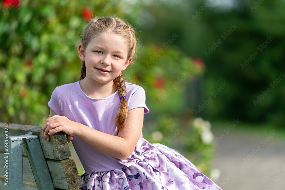 Fototapeta premium lächelndes, blondes Mädchen mit Zöpfen in lila Kleidung schaut an einem sonnigen Tag in die Kamera
