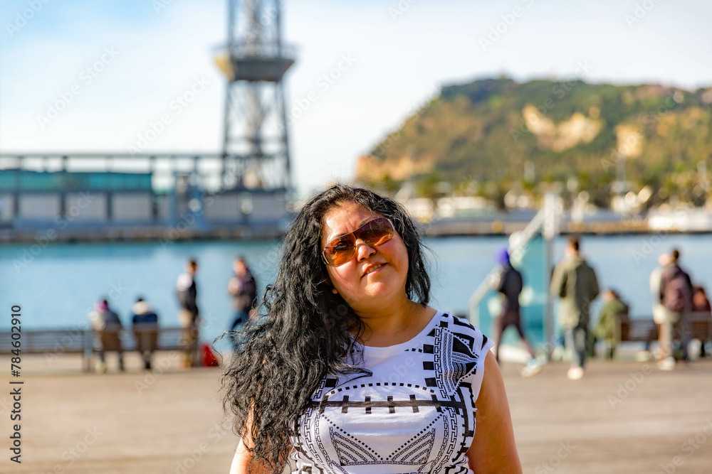 Waist up portrait of chubby Latin American woman on city promenade, cable car tower and mountain in blurred background, long black hair, sunglasses and looking at camera, sunny day in Barcelona, Spain