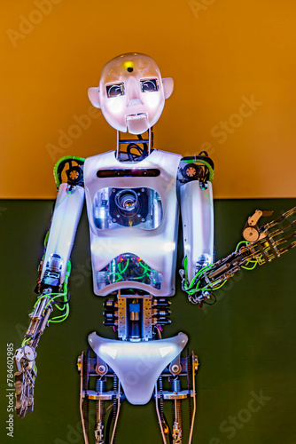 Front view of humanoid robot looking at camera, human face and body, cables and white lights, yellow and green wall in background. Concept of technology and automation in artificial intelligence