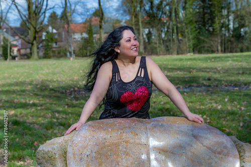 Smiling Latin American woman  in profile leaning on large stone, long black hair, blouse with heart, trees in blurred background, sunny day in Kasteelpark Elsloo park, South Limburg, Netherlands