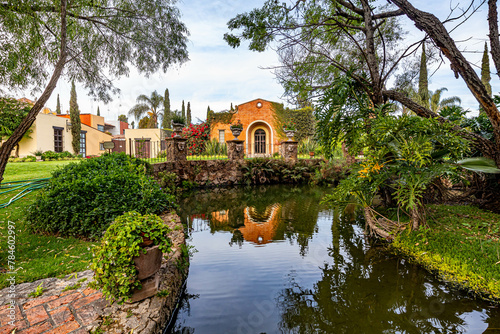 Garden of Mexican hacienda with small pond between trees and lush vegetation, colonial building in background, reflection in water surface, calm day in Zapopan, Jalisco Mexico