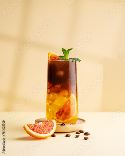 Americano coffee with orange juice (bumble) in a tall glass with ice and mint on a yellow background with coffee beans, shadows and citrus fruits. Trendy summer Asian refreshing drink.