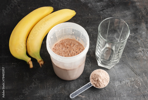 Blended chocolate protein drink in a shaker, plastic measuring spoon with protein powder, bananas and drinking glass on a dark background