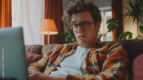 A perplexed young man in casual attire and glasses looks skeptically at his laptop screen while sitting on a couch photo